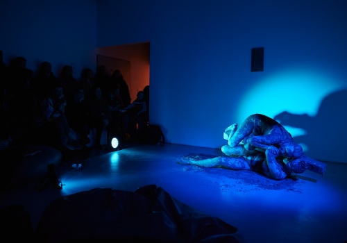 Zdjęcie. W sali wystawienniczej oświetlonej na niebiesko Monika Wińczyk i Magdalena Mellin wykonują performans. Artystki są ubrane w czarne kostiumy i wysmarowane gliną. W tle widać zebraną widownię, a na ścianie wisi praca artystyczna z wystawy.