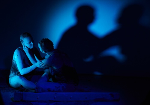 Zdjęcie. W sali wystawienniczej oświetlonej na niebiesko Monika Wińczyk i Magdalena Mellin wykonują performans. Artystki są ubrane w czarne kostiumy i wysmarowane gliną. Na ścianie widać ich cień.