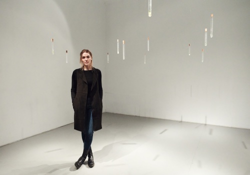 Zdjęcie. Portret cało postaciowy artystki Katarzyny Serkowskiej w jasnej sali wystawienniczej. Artystka stoi pomiędzy elementami instalacji artystycznej – nierównomiernie rozwieszonymi w przestrzeni sali probówkami. 