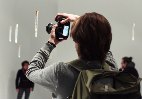 Zdjęcie. Osoba fotografująca aparatem fotograficznym instalację artystyczną składającą się z nierównomiernie rozwieszonych probówek. 