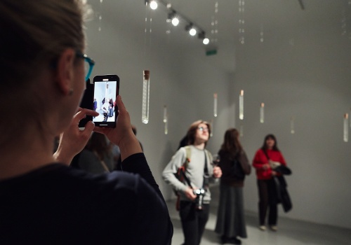 Zdjęcie. Publiczność oglądająca instalację artystyczną składającą się probówek. Osoba na pierwszym planie fotografuje probówki telefonem komórkowym. 