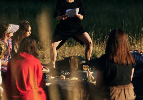 Zdjęcie. Ubrana na czarno kobieta, przodem do obiektywu czyta z kartki. Widoczne dwie kobiety od tyłu oraz dwie z boku. Wszystkie znajdują się na łące.