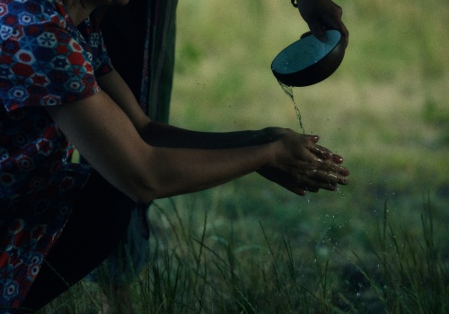 Zdjęcie. Kucającej kobiecie na łące polewane są ręce wodą z miseczki bambusowej przez drugą osobę, której widoczna jest tylko dłoń. 