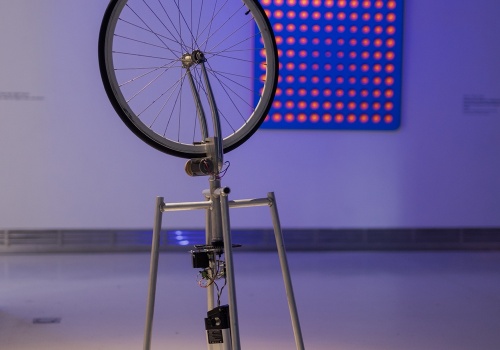 Zdjęcie. Na pierwszym planie praca Londona Tsai’a „Duchamp Cybernetyczny” składająca się z elementów składowych roweru. W tle obraz Wen-Yinga Tsai’a  „Nałożony obraz: niebieski i pomarańczowy”.