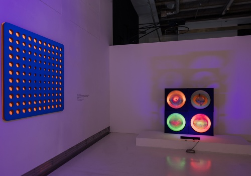 Zdjęcie. Przestrzeń wystawy z widocznymi dwiema pracami Wen-Yinga Tsai’a wykorzystującymi światło ultrafioletowe. Po lewej obraz „Nałożony obraz: niebieski i pomarańczowy”, po prawej „Ściana Multi-kinetyczna” w ruchu.