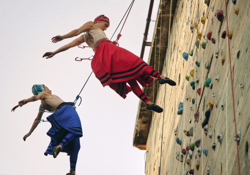 Zdjęcie. Dwie kobiety ubrane w kolorowe stroje inspirowane tradycją kaszubską wykonujące wyskok z asekuracją, na ściance wspinaczkowej. 