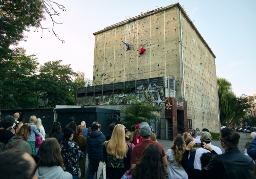 Zdjęcie. Grupa publiczności obserwująca budynek klubu Bunkier, na którego ściance wspinaczkowej znajdują się dwie kobiety ubrane w kolorowe stroje inspirowane tradycją kaszubską.