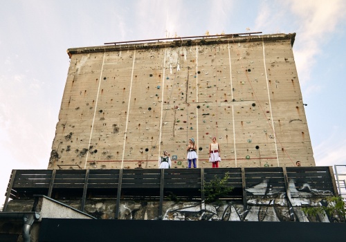 Zdjęcie. Na tle ścianki wspinaczkowej klubu Bunkier w Gdańsku, za balustradą, stoją trzy kobiety ubrane w kolorowe stroje inspirowane tradycją kaszubską oraz dwóch mężczyzn w ciemnych koszulkach.