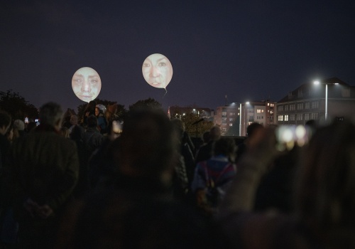 Na zdjęciu widok na tłum osób znajdujących się na dachu Gdańskiego Teatru Szekspirowskiego. Osoby pokazane od tyłu w trakcie oglądania powietrznego performansu - projekcji twarzy na ponad dwumetrowych balonach, znajdujących się ponad ich głowami. W tle panorama Gdańska i nocne niebo.