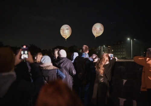 Na zdjęciu widok na tłum osób znajdujących się na dachu Gdańskiego Teatru Szekspirowskiego. Osoby pokazane od tyłu w trakcie oglądania powietrznego performansu - projekcji twarzy na ponad dwumetrowych balonach, znajdujących się ponad ich głowami. Osoby rozświetlone pobliskimi reflektorami. W tle panorama Gdańska i nocne niebo.