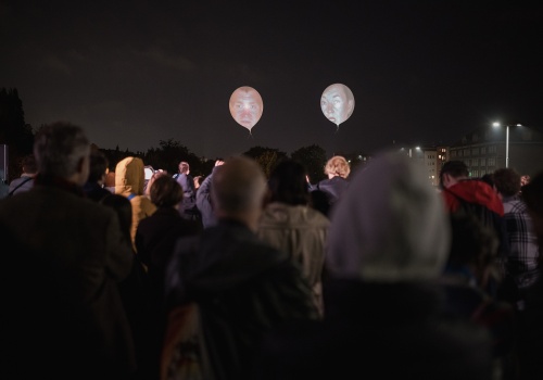 Na zdjęciu widok na tłum osób znajdujących się na dachu Gdańskiego Teatru Szekspirowskiego. Osoby pokazane od tyłu w trakcie oglądania powietrznego performansu - projekcji twarzy na ponad dwumetrowych balonach, znajdujących się ponad ich głowami. W tle panorama Gdańska i nocne niebo.