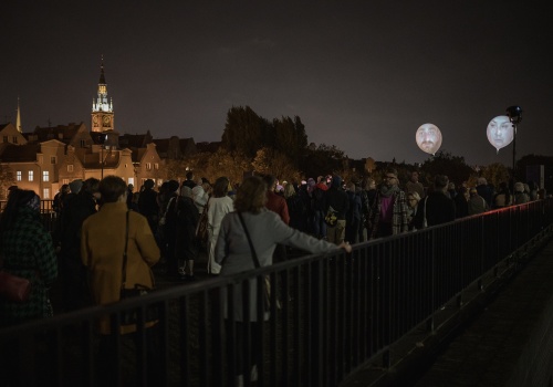 Na zdjęciu widok na tłum osób znajdujących się na dachu Gdańskiego Teatru Szekspirowskiego, widoczna barierka tarasu. Osoby pokazane od tyłu w trakcie oglądania powietrznego performansu - projekcji twarzy na ponad dwumetrowych balonach, znajdujących się ponad ich głowami. W tle panorama Gdańska i nocne niebo.