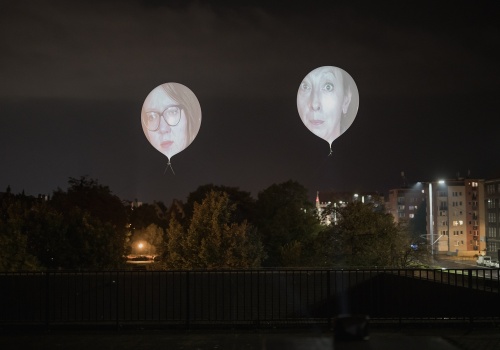 Zbliżenie na powietrzny performans - dwa ponad dwumetrowe balony na których wyświetlone zostały twarze kobiet, uczestniczek projektu. Widoczny również teren dachu Gdańskiego Teatru Szekspirowskiego. W tle widok na panoramę Gdańska.