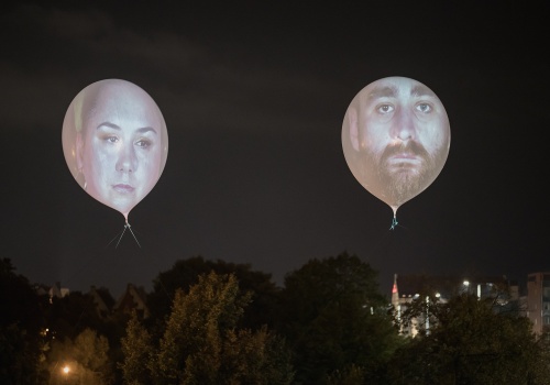 Zbliżenie na dwa ponad dwumetrowe balony na których wyświetlone zostały twarz kobiety i mężczyzny, uczestników projektu. W tle widok na nocne niebo i na panoramę Gdańska. 