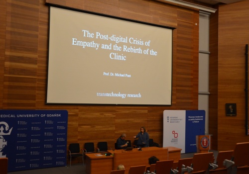 Zdjęcie. Na Sali wykładowej znajduje się mężczyzna oraz kobieta. W centralnej części widać ekran, na którym wyświetlany jest slajd z napisem: The Post-Digital Crisis of Empathy and Rebirth of the Clinic.