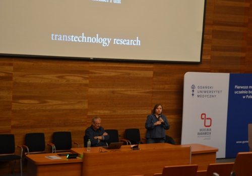 Zdjęcie. Na Sali wykładowej znajduje się mężczyzna oraz kobieta. Kobieta mówi przez mikrofo.n W centralnej części widać ekran, na którym widoczne są napisy transtechnology research.