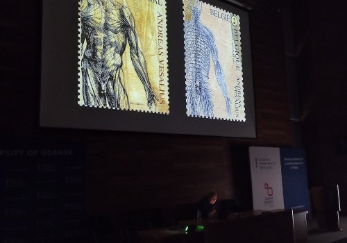 Zdjęcie. W przyciemnionej Sali wykładowej wykładowca wygłasza wykład. Widoczny jest ekran ze slajdami dotyczącymi anatomii ludzkiego ciała.