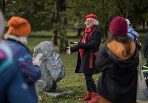 Prowadzący spacer Andrzej Stelmasiewicz wskazuje rzeźbę z kamienia przedstawiającą profil Jana Heweliusza. Wokół widać ludzi, którzy przyglądają się rzeźbie i prowadzącemu spacer.