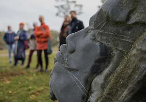 Na pierwszym planie kamienna rzeźba przedstawiająca profil Jana Heweliusza. Za nią w oddali widać rozmazane sylwetki osób, które oglądają rzeźbę.