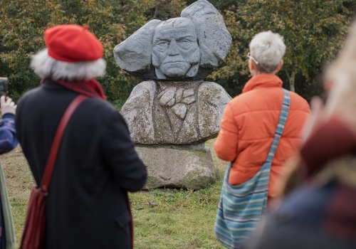 Kilka osób stoi i przygląda się widocznej na drugim planie kamiennej rzeźbie przedstawiającej Artura Schopenhauera. Za rzeźbą widać drzewa i zieleń Parku Reagana.