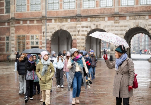 Na pierwszym planie przewodniczka prowadząca wycieczkę, koordynatorka projektu GAPS. Kobieta w prawej ręce trzyma parasol rozłożony nad głową, spogląda również w prawo, na grupę osób podążającą za nią. Widzimy opiekunkę grupy oraz kilkudziesięcioro nastoletnich uczniów, podążających za przewodniczką. W tle widać budynek Zielonej Bramy w Gdańsku.