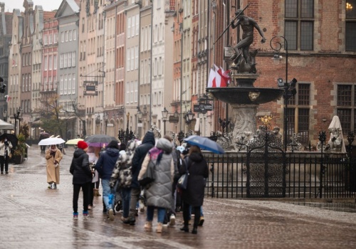 Grupa osób pod parasolkami przechodzi obok Fontanny Neptuna na Długim Targu w Gdańsku. Osoby są zwrócone tyłem do obiektywu, podążają w kierunku Ratusza Głównomiejskiego. 