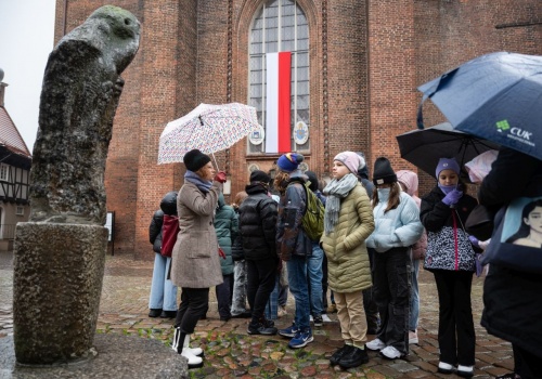 Od lewej strony widoczna abstrakcyjna rzeźba z kamienia, kształtem przypominająca skulonego siedzącego na gałęzi ptaka. Tuż obok rzeźby widzimy, stojącą bokiem przewodniczkę spaceru, która mówi do grupy młodzieży spoglądającej na rzeźbę, ale i na znajdującą się za nimi bazylikę Mariacką. W tle widoczny jest fragment fasady Bazyliki Mariackiej, z wywieszoną polską flagą.