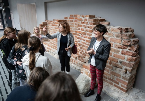 Grupa młodzieży znajduje się we wnętrzu nowoczesnego budynku, widoczna od tyłu, patrzy na dwie kobiety, które mówią do nich. Za kobietami tuż przy szarej ścianie budynku widoczny jest fragment zabytkowego, ceglanego muru.