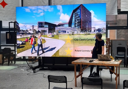 Zdjęcie. Na przestrzeni Patio znajduje się kobieta. W centralnej części widać ekran, na którym wyświetlany jest slajd z napisem: City Centre Campus.