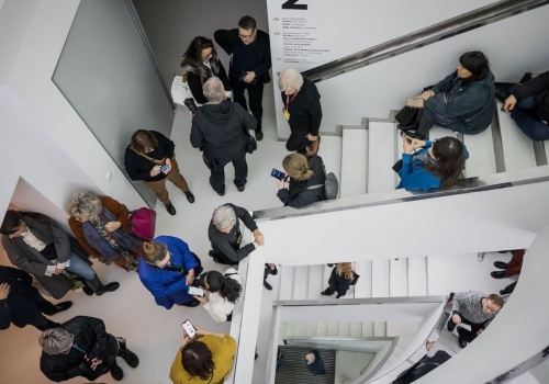 Na fotografii widoczny tłum osób idących klatką schodową w stronę piętra z wystawą „Spragnieni miasta”. Widok z góry na dusze klatki schodowej. Część osób rozmawia z innymi, część przygląda się smartfonom, część opiera się o rampę schodów, oczekując na rozpoczęcie wydarzenia.