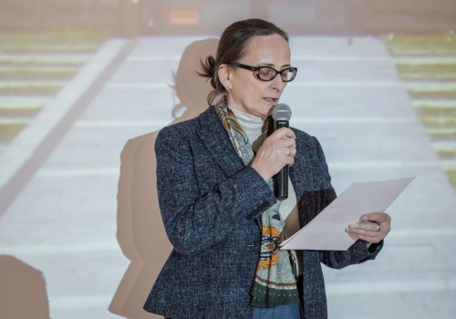 Dyrektor CSW ŁAŹNIA Jadwiga Charzyńska podczas oficjalnego otwarcia wystawy „spragnieni miasta”, podczas oficjalnej mowy i otwarcia wystawy. W tle widoczne wyświetlone z rzutnika zdjęcie przedstawiające ulicę i wejście do budynku.