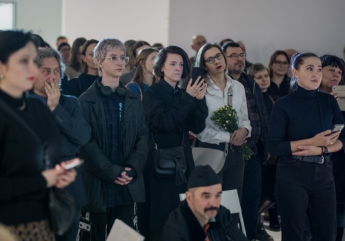 Widoczny tłum ludzi słuchających mów otwierających wernisaż wystawy „spragnieni miasta”. Niektóre osoby trzymają w dłoniach kwiaty dla artysty, część osób ma w dłoniach smartfony, którymi dokumentowali wydarzenie.
