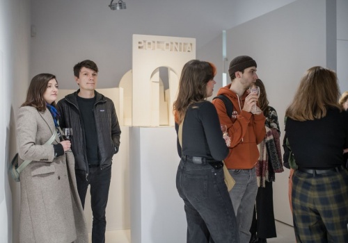 Grupa ludzi otaczająca model pawilonu polskiego na Biennale w Wenecji projektu prof. Jacka Dominiczaka. Na modelu jest wyraźnie wyróżniający się napis „POLONIA”, umieszczony centralnie nad zaznaczonym wejściem.