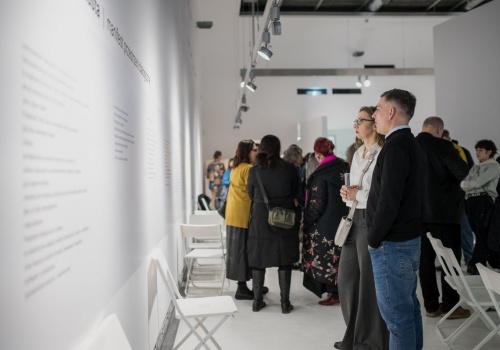Tłum ludzi przygląda się ścianie na ekspozycji z tekstem manifestu architektury dialogicznej, między ludźmi widoczne są białe krzesełka, będące częścią aranżacji wystawy. W tle widać pozostałą część sali ekspozycyjnej oraz ścianę „diaformy”.