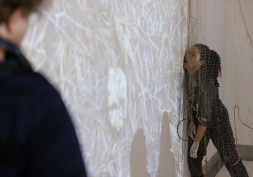 Zdjęcie. Performerka Dominika Kimaty stoi w sali wystawienniczej oparta policzkiem o ścianę.