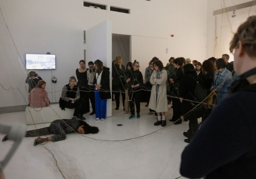 Zdjęcie. Performerka Dominika Kimaty leży na podłodze sali wystawienniczej. W okół niej znajdują się prace prezentowane na wystawie Pameli Leończyk oraz zgromadzona publiczność.
