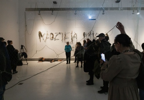 Zdjęcie. Performerka Dominika Kimaty leży na podłodze sali wystawienniczej. Wokół niej znajdują się prace prezentowane na wystawie Pameli Leończyk oraz zgromadzona publiczność.