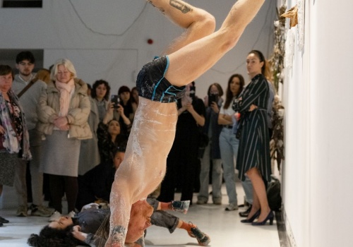 Zdjęcie. Performer Łukasz Wójcicki stoi na rękach, opierając się jedną nogą o ścianę sali wystawienniczej. W tle znajduje się performerka Dominika Kimaty leżąca na podłodze oraz zgromadzona publiczność.