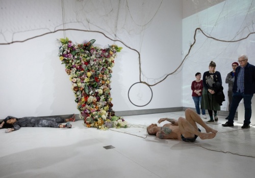 Zdjęcie. Performans Dominiki Kimaty i Łukasz Wójcickiego podczas wernisażu wystawy Pameli Leończyk w CSW ŁAŹNIA. Kobieta i mężczyzna leżą na podłodze. Obok nich znajduje się praca artystyczna – tren z kwiatów oraz zgromadzona publiczność.