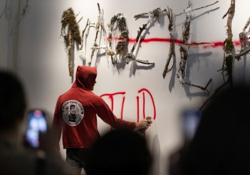 Zdjęcie. Performer Łukasz Wójcicki maluje spray’em czerwone kreski i litery na pracy artystycznej Pameli Leończyk umieszczonej na ścianie sali wystawienniczej.